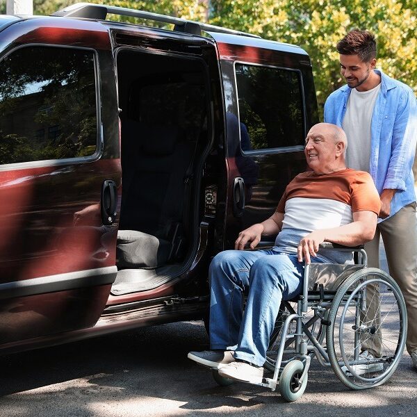 Young man helping patient in wheelchair to get into van outdoors